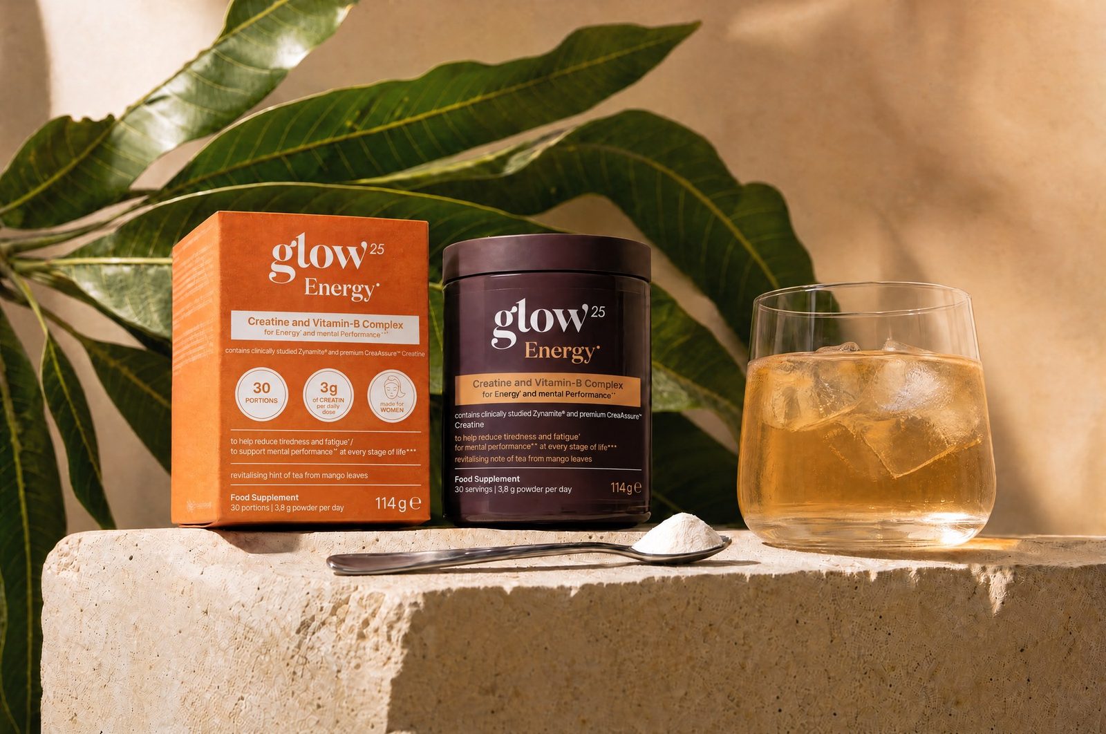 Confezione e barattolo di Glow25 Energy® su travertino, accanto foglie di mango, cucchiaino e un bicchiere con una bevanda ghiacciata