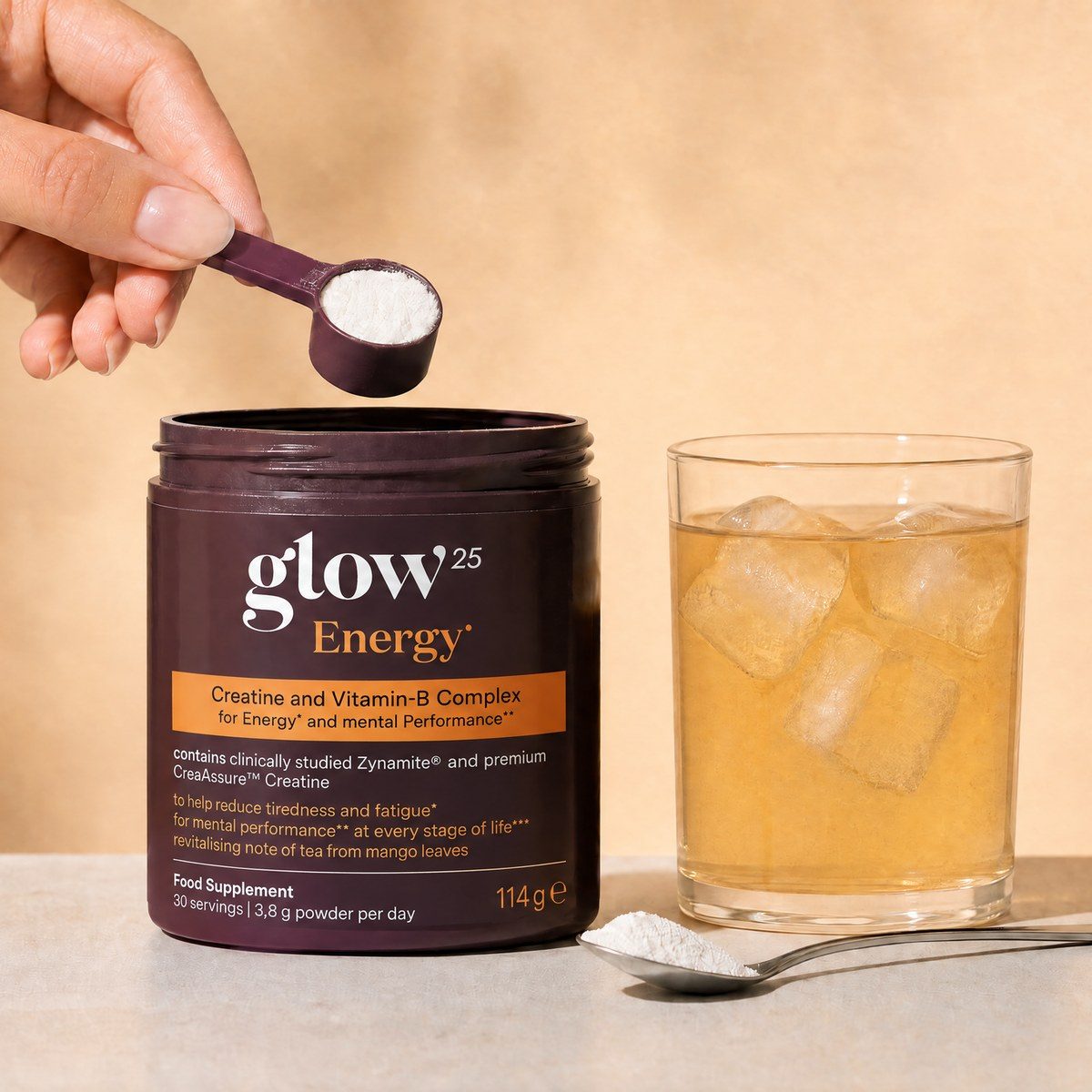 Una mano solleva il misurino dal barattolo di Glow25 Energy, accanto un cucchiaino con la polvere e un bicchiere con una bevanda ghiacciata